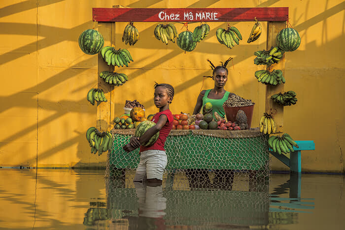 ボードワン・ムワンダ（Baudouin Mouanda）<br />
 ＜シリーズタイトル＞ 『ある季節の空』<br />
（Ciel de saison / Seasonal Sky）<br />
＜作品タイトル＞ 野菜売りの店で（2020年）<br />
（Chez la marchande de légumes）<br />
▶コロナ禍にあった2020年にコンゴ共和国の首都ブラザビルで起きた洪水を、被災者たちの協力で再現した。現地では雨の脅威が恒常的なものになっている<br />
Credit: Courtesy Baudouin Mouanda
